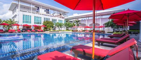 A swimming pool with red lounge chairs and umbrellas, surrounded by a modern building with large windows.