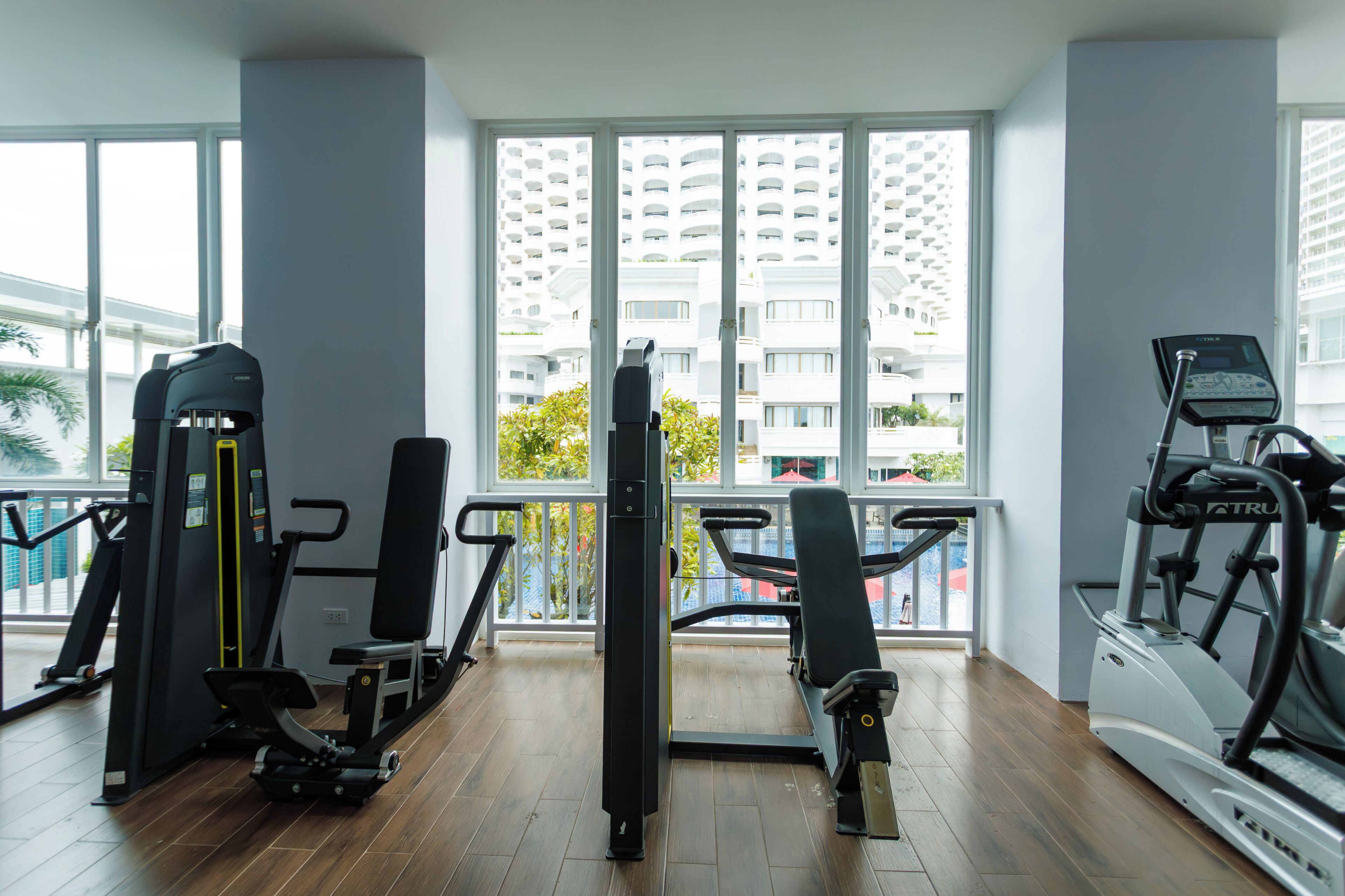 Un gimnasio con máquinas de cardio, máquinas de pesas y vistas a un edificio alto a través de amplias ventanas.