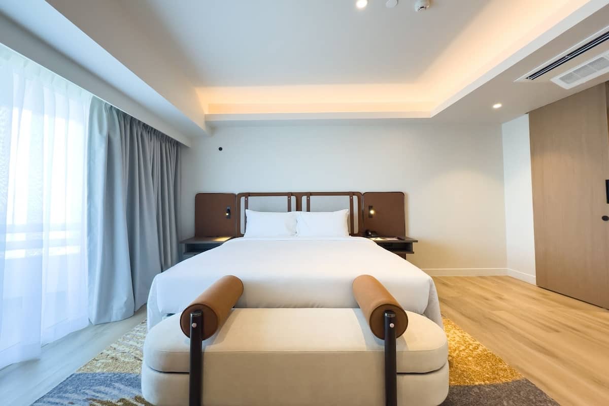Une chambre d’hôtel moderne avec un grand lit, une table de chevet, une commode, une porte en bois et une grande fenêtre avec des rideaux.