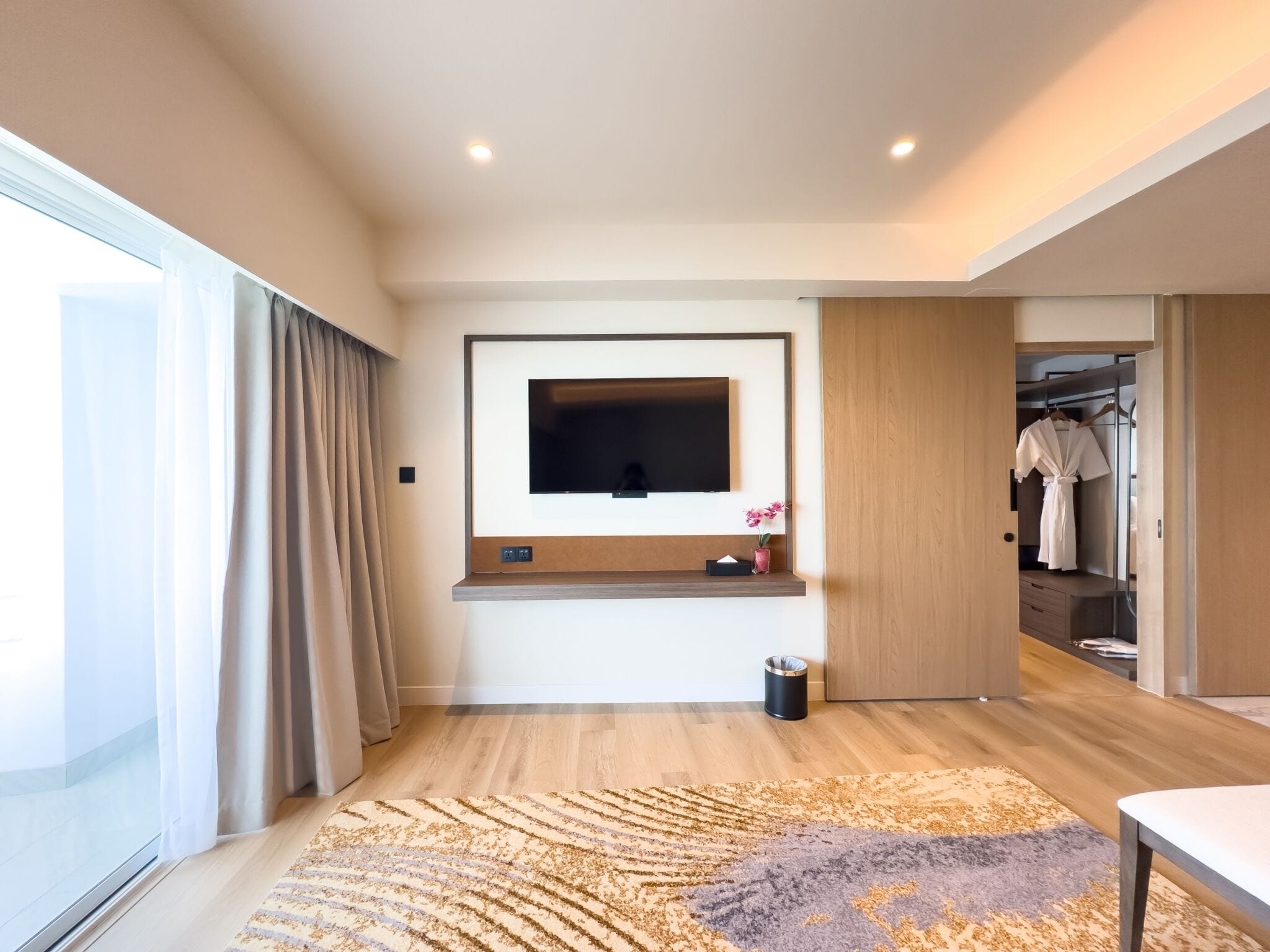A modern bedroom with a large TV mounted on the wall, a wooden floor, a patterned rug, and a walk-in closet.