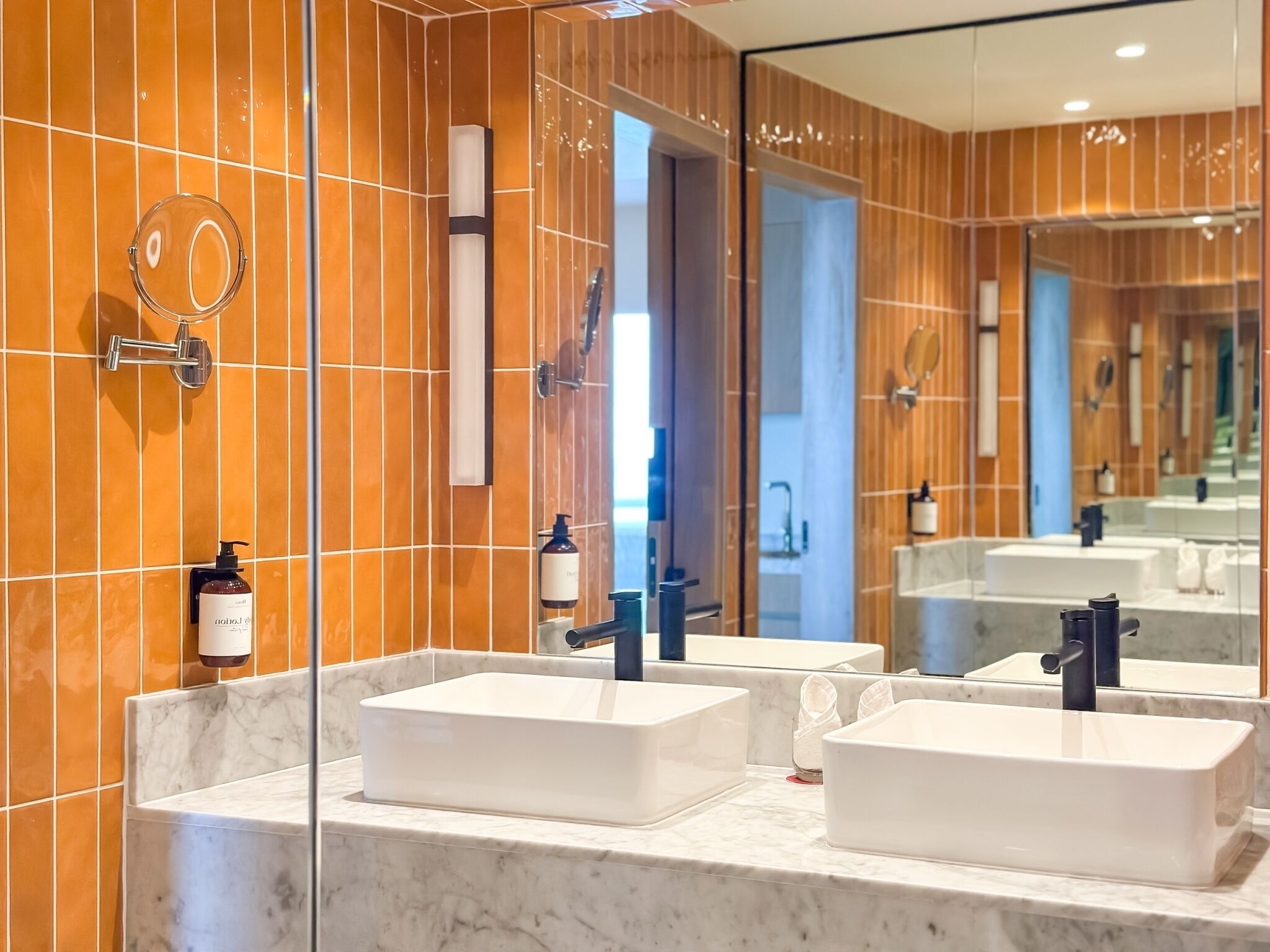 A modern bathroom with two white sinks, a large mirror, and orange-tiled walls.
