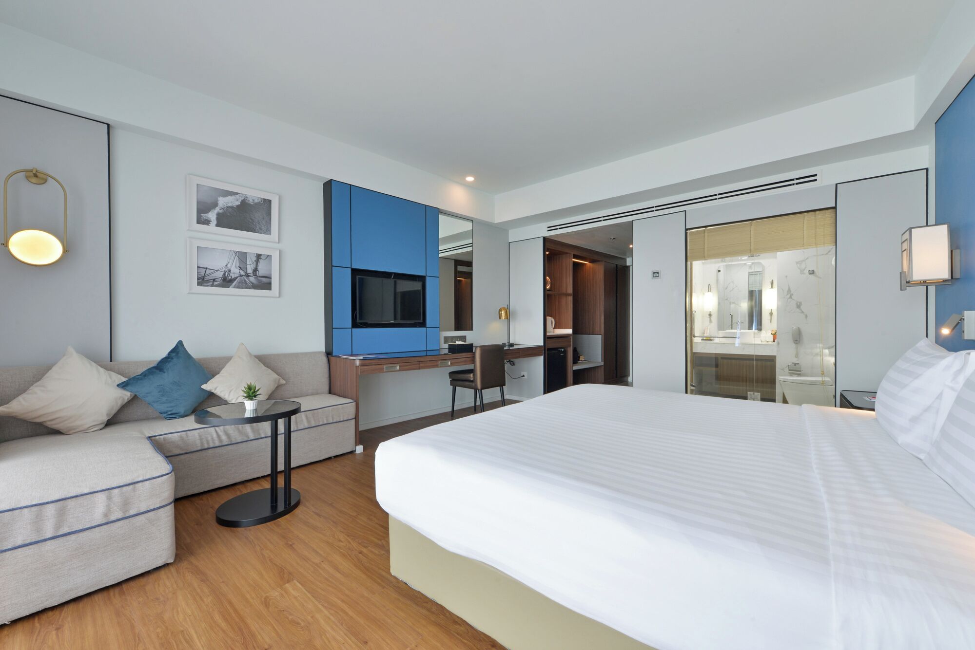 A modern hotel room with a large bed, a sofa, a desk with a TV, and a bathroom visible through a glass partition.