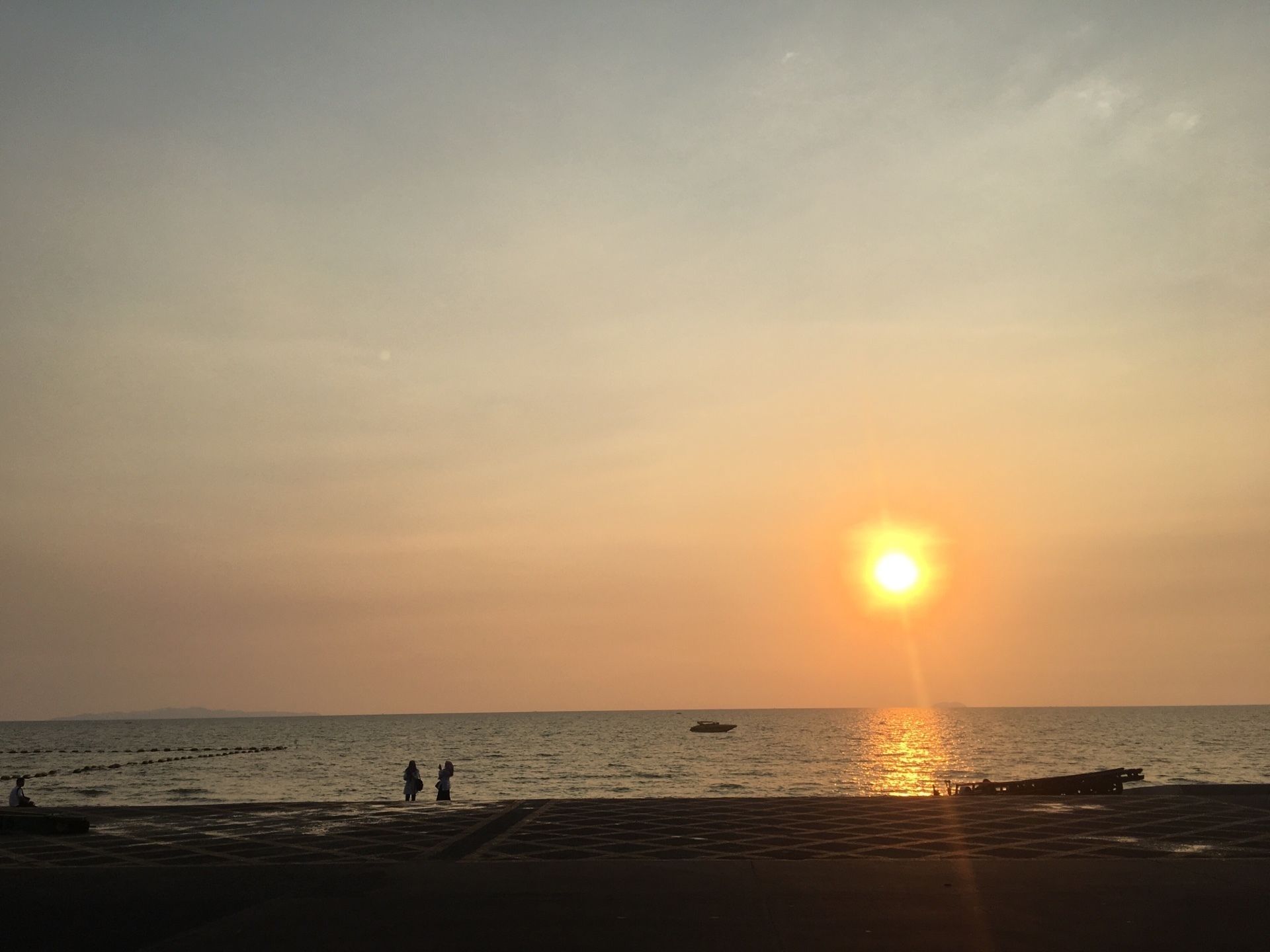 A beach at sunset with a clear sky, calm sea, and a few people walking along the shore.
