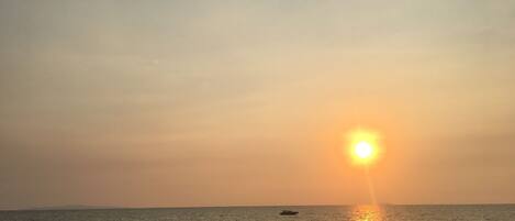 A beach at sunset with a clear sky, calm sea, and a few people walking along the shore.