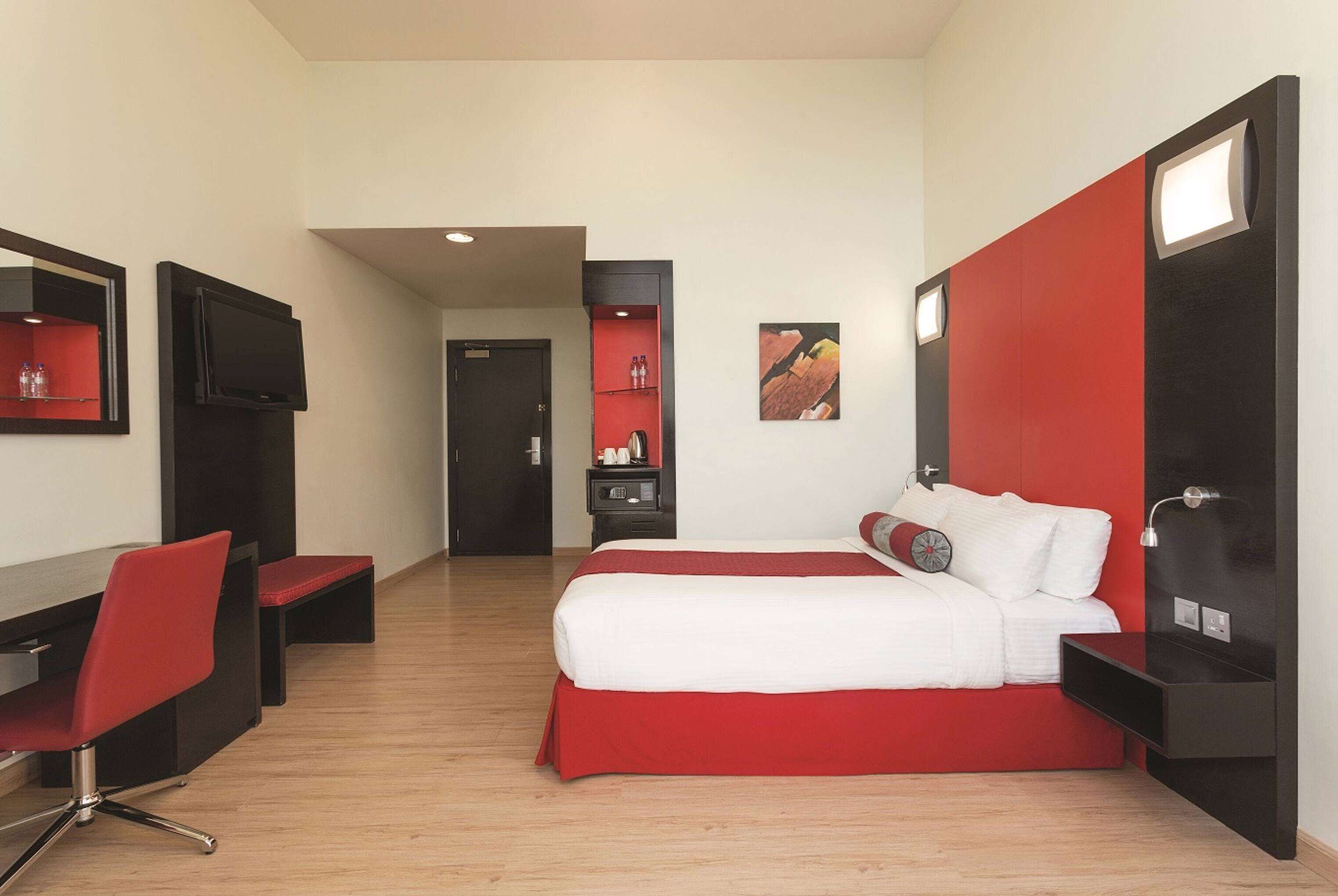 Habitación de hotel con cama, escritorio, silla, televisor y una pared de acento roja.
