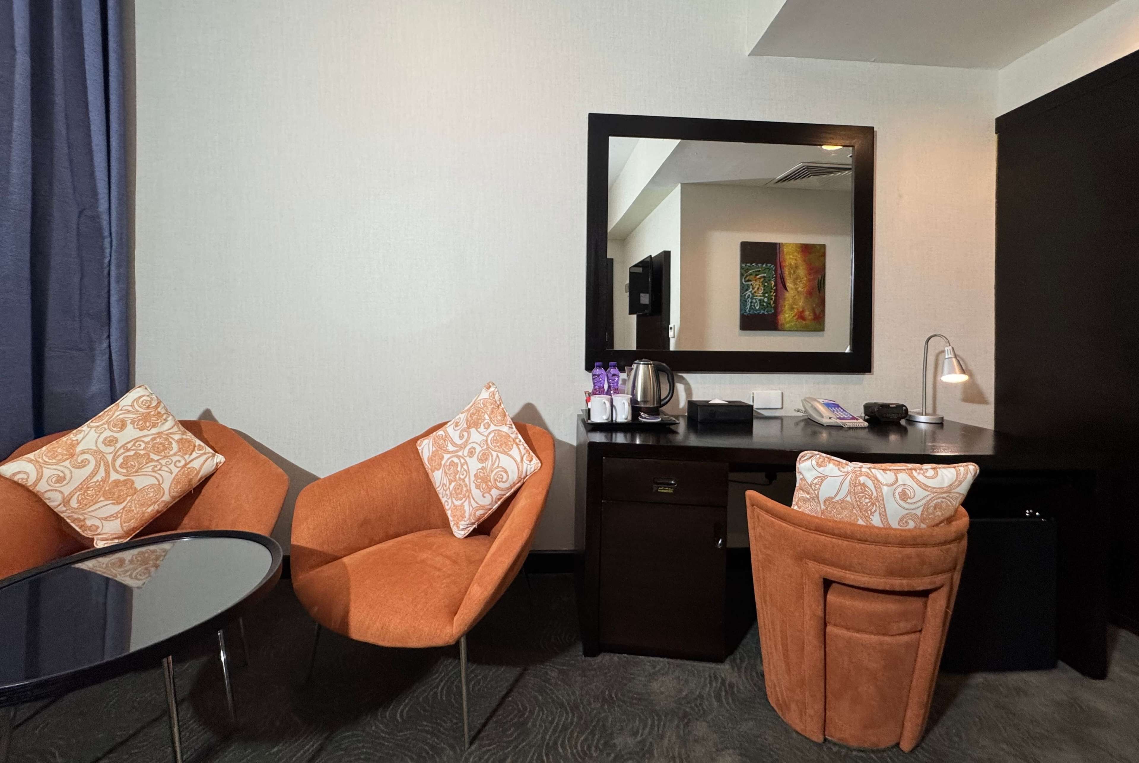 A hotel room with two orange chairs, a small table, a desk with a mirror, a coffee maker, and a lamp.