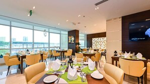 A modern dining area with multiple tables set for meals, large windows offering a city view, and a flat-screen TV mounted on the wall. - Ramada Encore by Wyndham Doha (Doha)