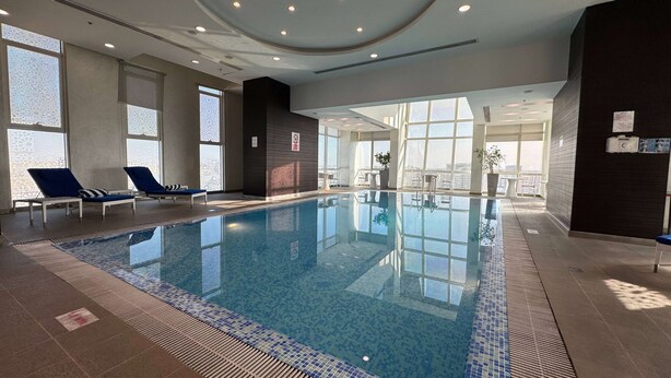 An indoor swimming pool with lounge chairs, large windows, and a view of the city. - Ramada Encore by Wyndham Doha (Doha)