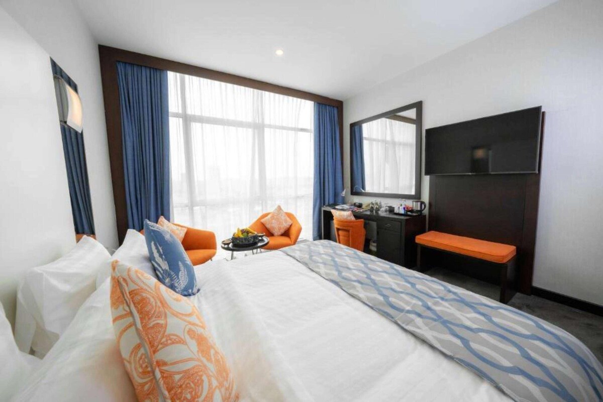 Habitación de hotel con una cama grande, un escritorio con silla, un banco, un televisor y una ventana con cortinas.
