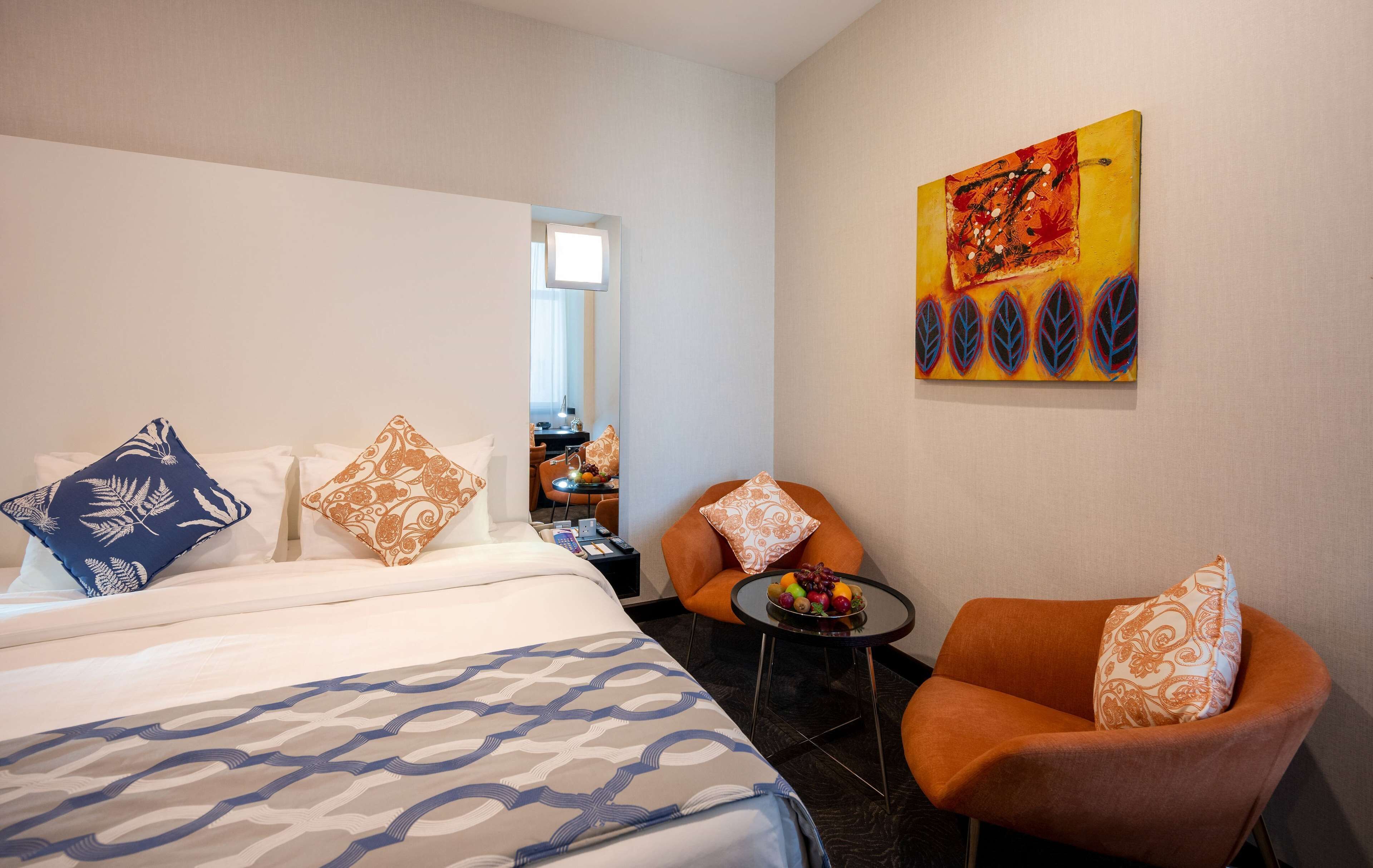 Habitación de hotel con cama, dos sillas, una mesita con fruta y un cuadro en la pared.