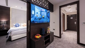 A hotel room with a large flat-screen TV mounted on a dark wooden console, a bed with white bedding, a fruit bowl on the console, and a door leading to another room. - Ramada Encore by Wyndham Doha (Doha)