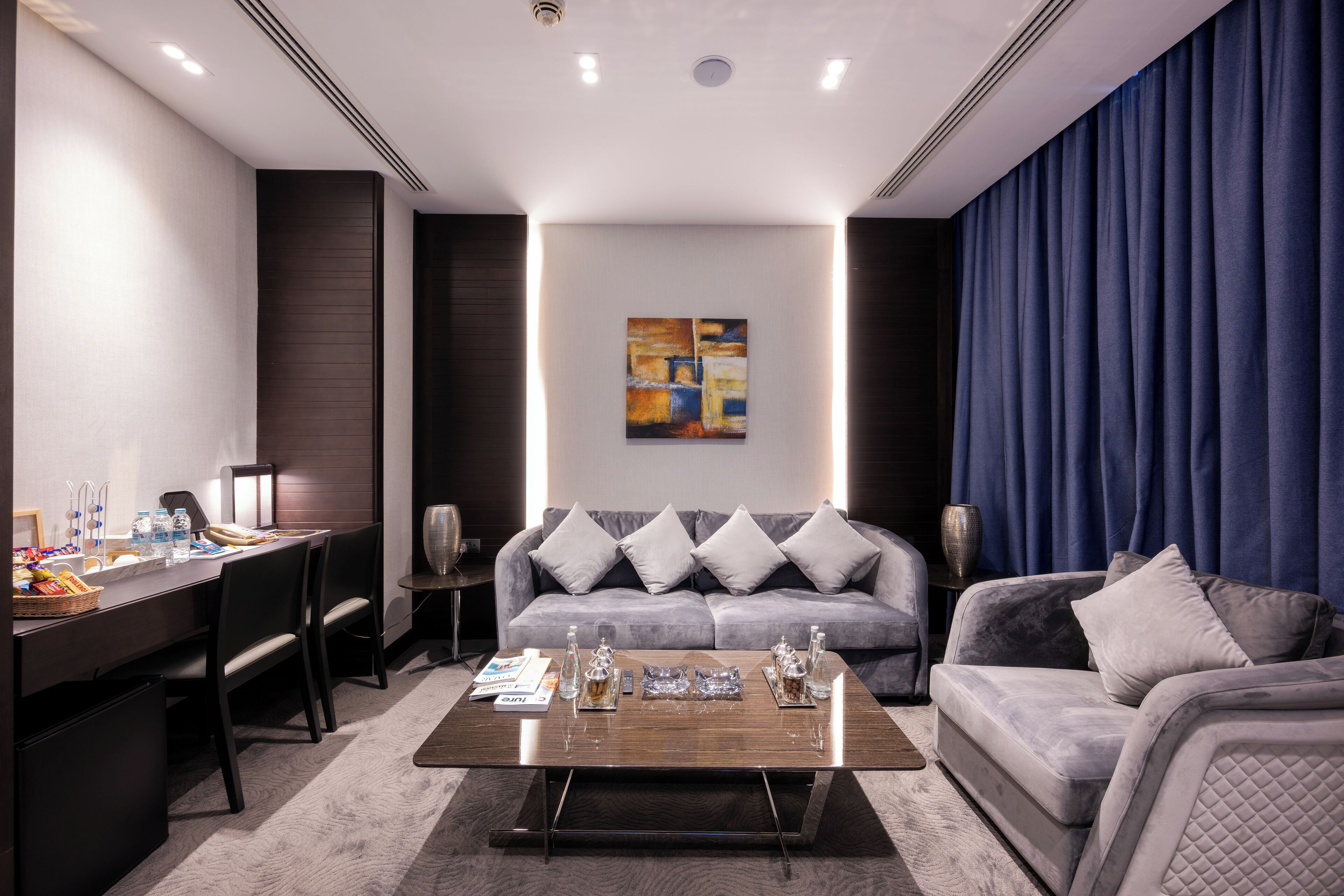 A modern hotel room with a sofa, a coffee table, a desk, and a painting on the wall.