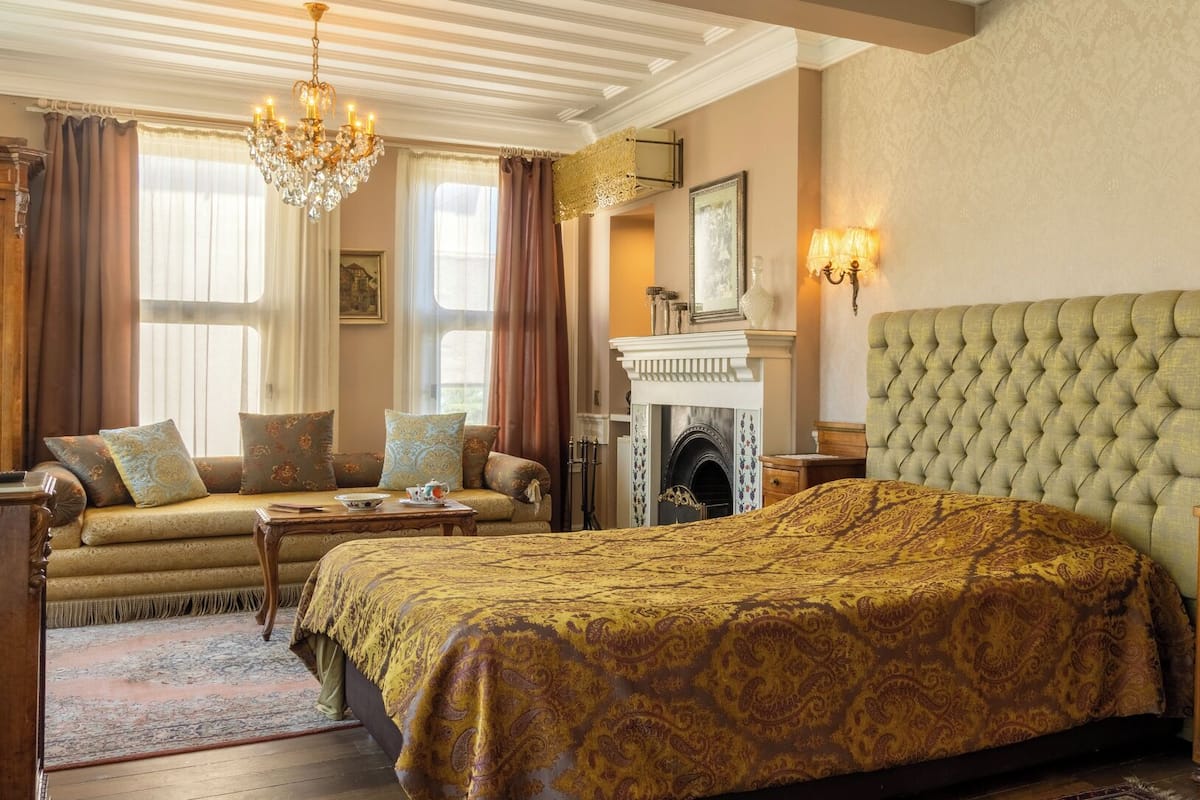 A traditional room with a large bed, a sofa, a fireplace, and a chandelier.