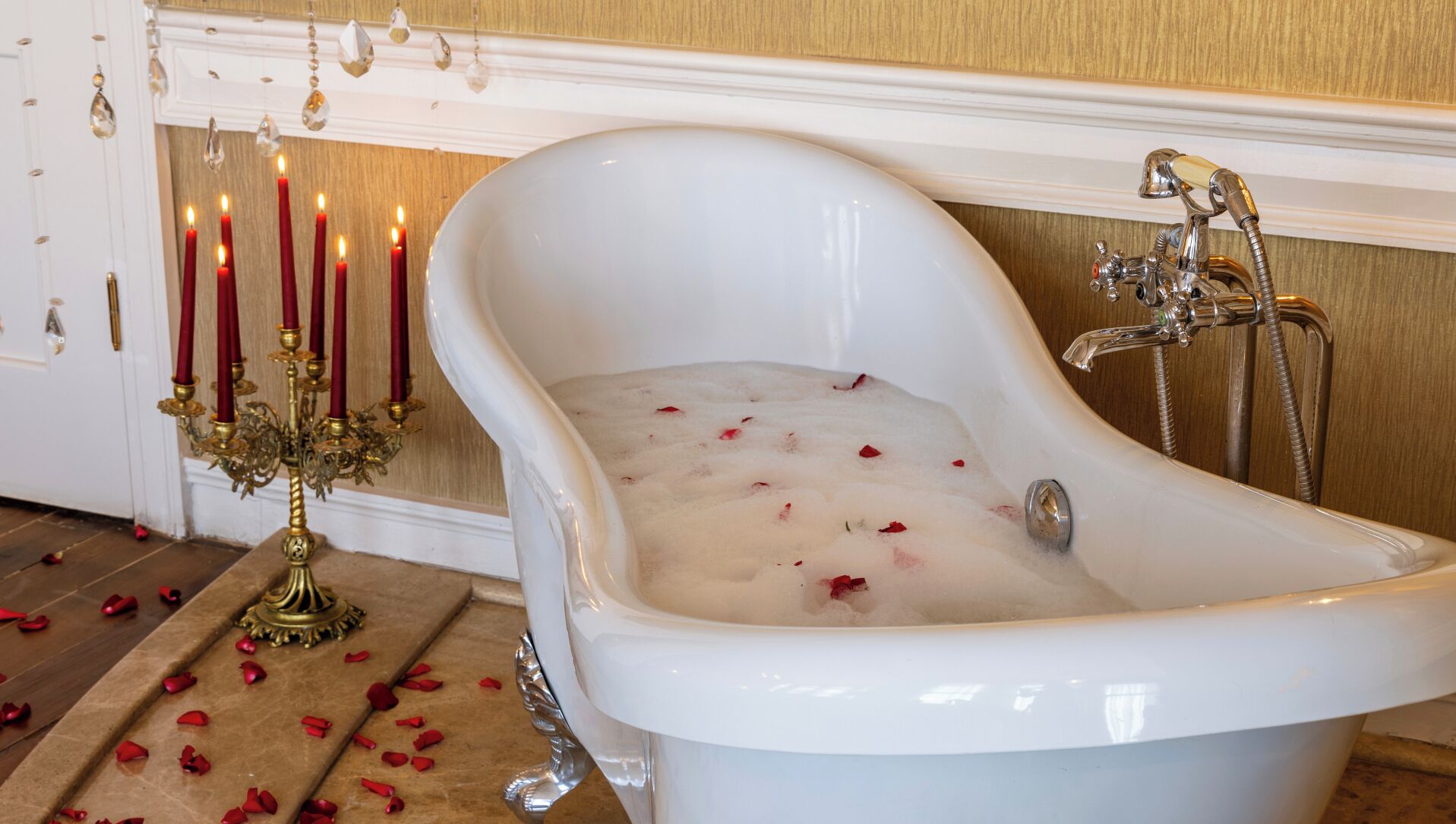 Una bañera blanca tipo pata de león con pétalos de rosa en el suelo, un candelabro dorado con velas rojas, y una ducha con grifo antiguo.