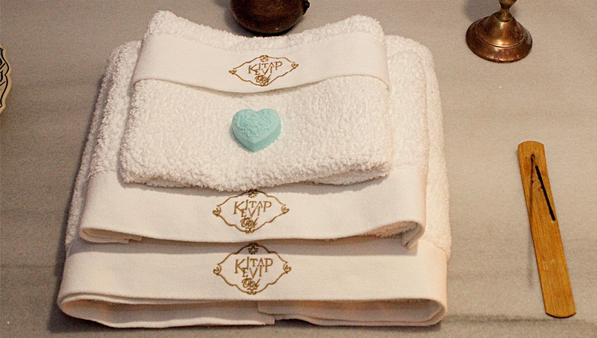 Una toalla blanca doblada con pulcritud, con un objeto con forma de corazón y el texto 'KITAP EVI' bordado, junto a un objeto de madera y dos objetos metálicos.