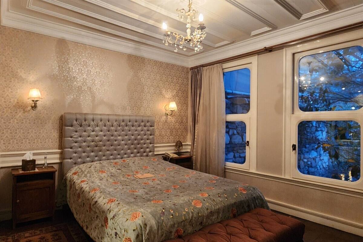 A bedroom with a tufted headboard, floral bedspread, two bedside lamps, a nightstand, and a window with a view of trees.
