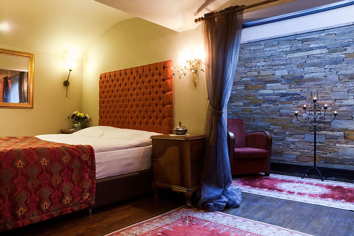 A bedroom with a tufted headboard, a large bed, a nightstand with a vase, a red chair, a stone wall, and a floor lamp.