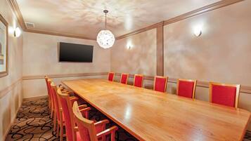 A conference room with a long wooden table, red upholstered chairs, a mounted flat-screen TV, and a patterned carpet.