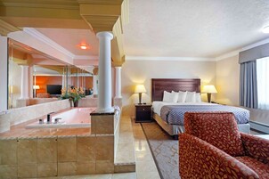 Camera d'albergo con un letto grande, due lampade da comodino, una poltrona rossa con motivo decorativo, un tavolino, un comodino, uno specchio grande e vista sulla zona della hall.