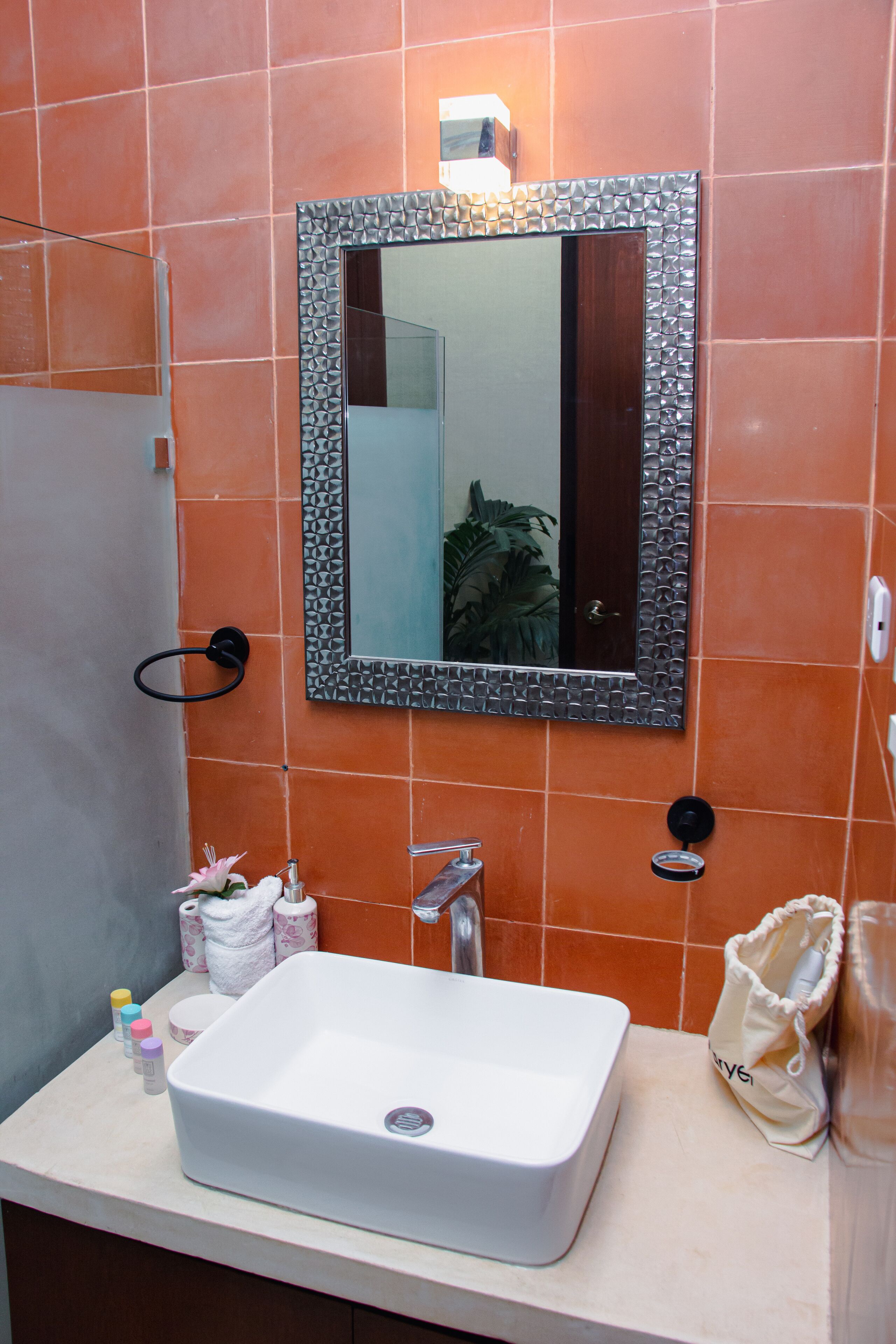 Une salle de bain avec un lavabo blanc, un miroir doté d’un cadre texturé, des carreaux orange, un porte-serviettes et une petite plante.