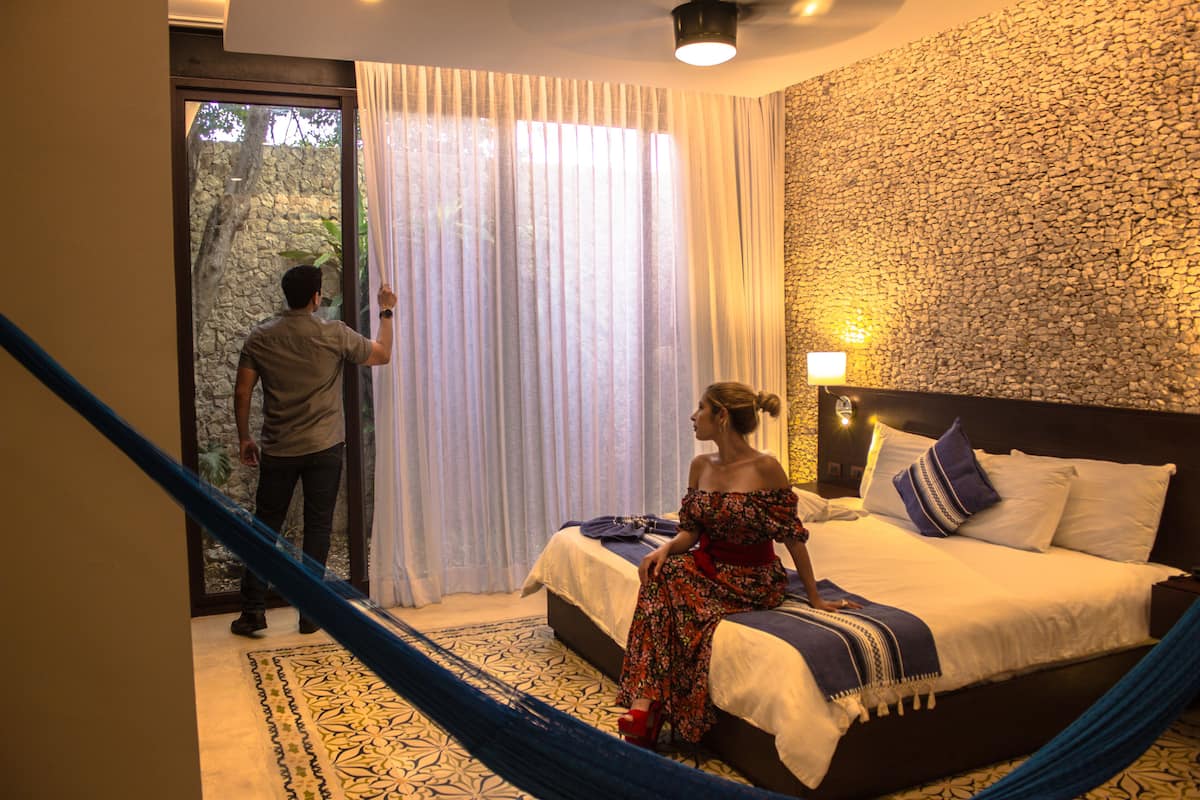 A bedroom with a bed, a large window with curtains, a stone wall, and a hammock.
