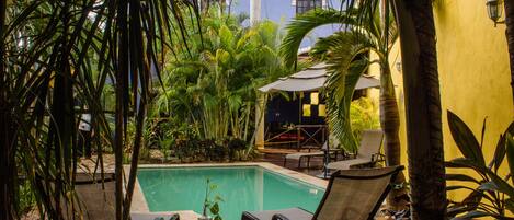 Área de piscina com espreguiçadeiras e uma mesa, cercada por plantas tropicais e um edifício ao fundo.