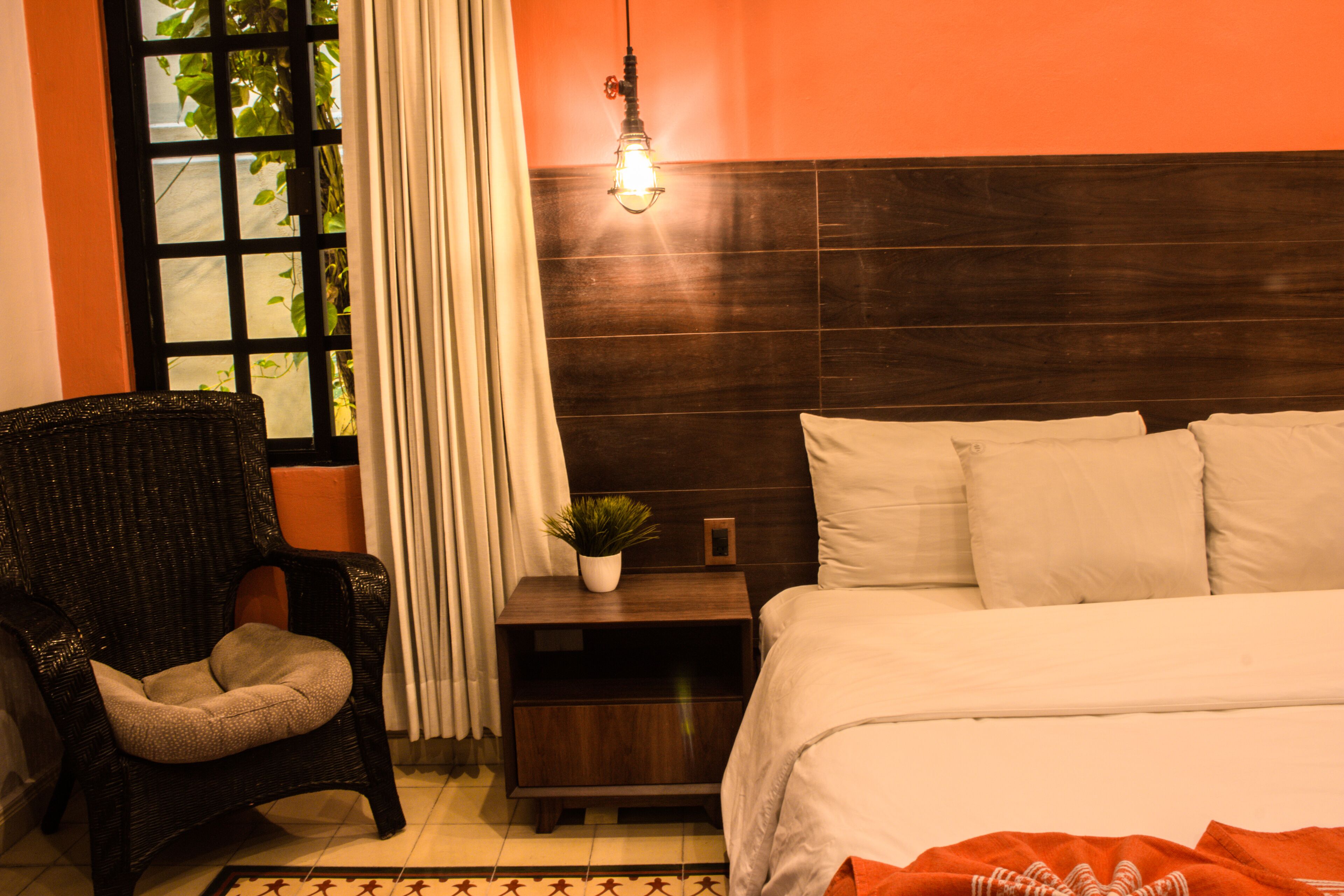 A bedroom with a bed, a nightstand, a chair, a window with curtains, and a hanging light fixture.