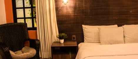 A bedroom with a bed, a nightstand, a chair, a window with curtains, and a hanging light fixture.