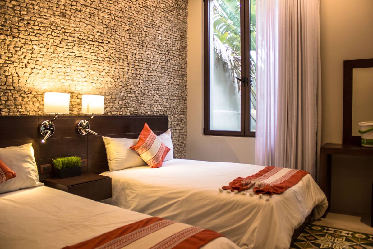 A hotel room with two beds, a stone wall, a window with curtains, and a nightstand with a lamp.
