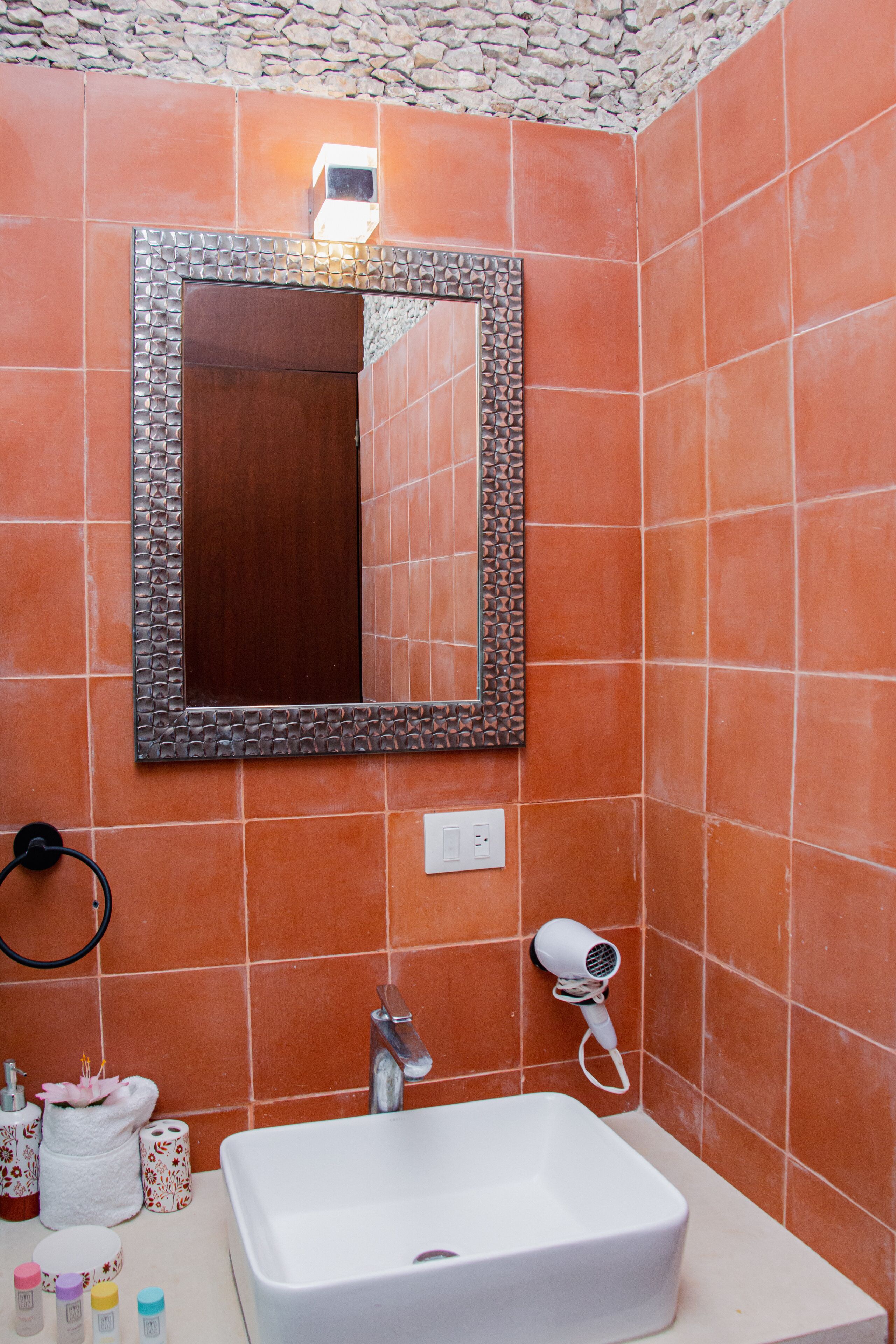 Une salle de bain avec un lavabo blanc, un miroir doté d’un cadre décoratif, un mur recouvert de carreaux orange, un sèche-cheveux et des articles de toilette posés sur le comptoir.