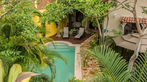 Une piscine entourée d’une végétation luxuriante et un bâtiment jaune avec un toit recouvert de tuiles.