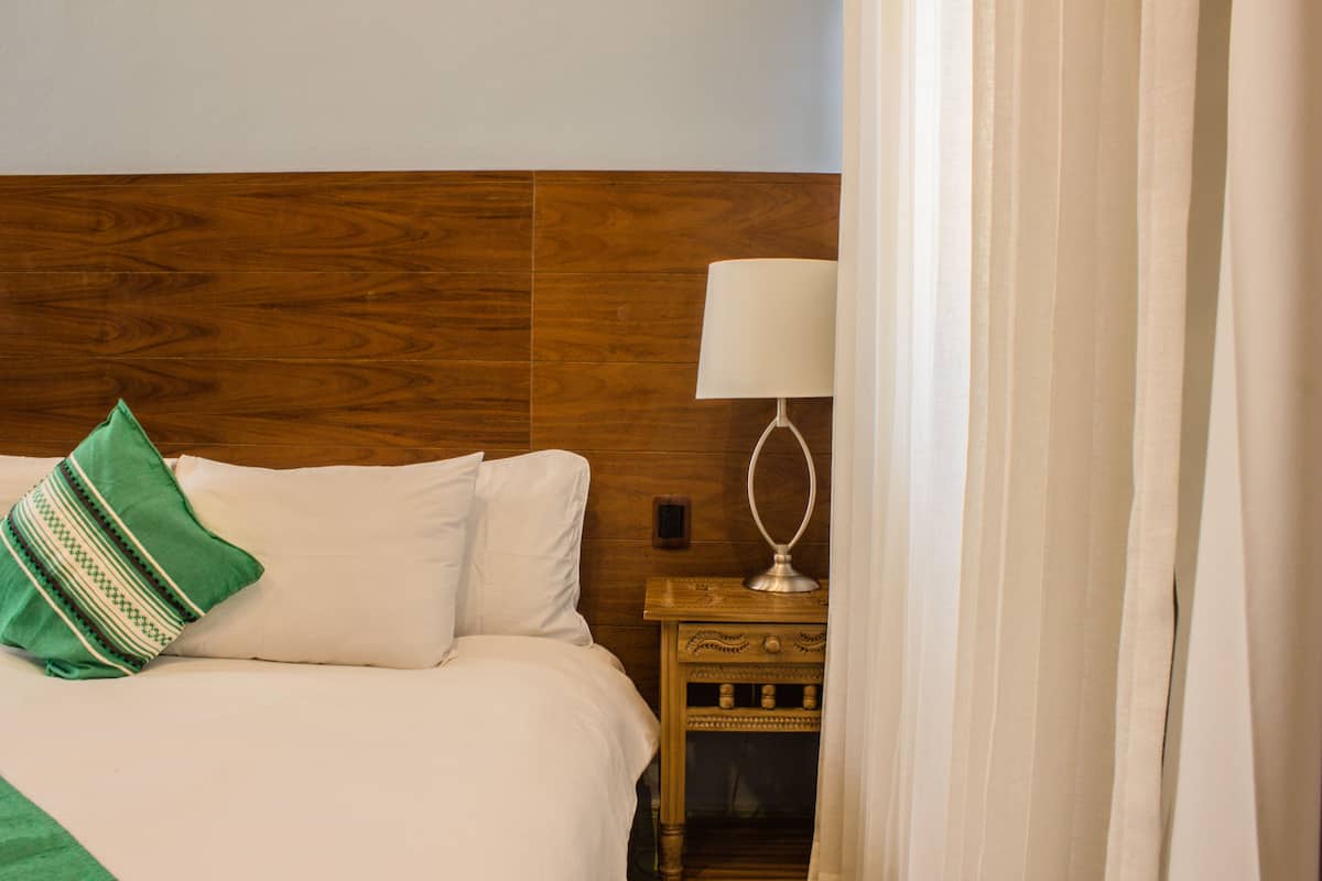 A hotel room with a wooden headboard, a bed with white linens, a green and white pillow, a bedside table with a lamp, and a curtain partially drawn.