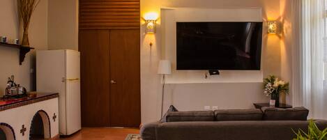 Una sala de estar con sofá, televisión montada en la pared, una puerta de madera y una pequeña cocina con refrigerador.