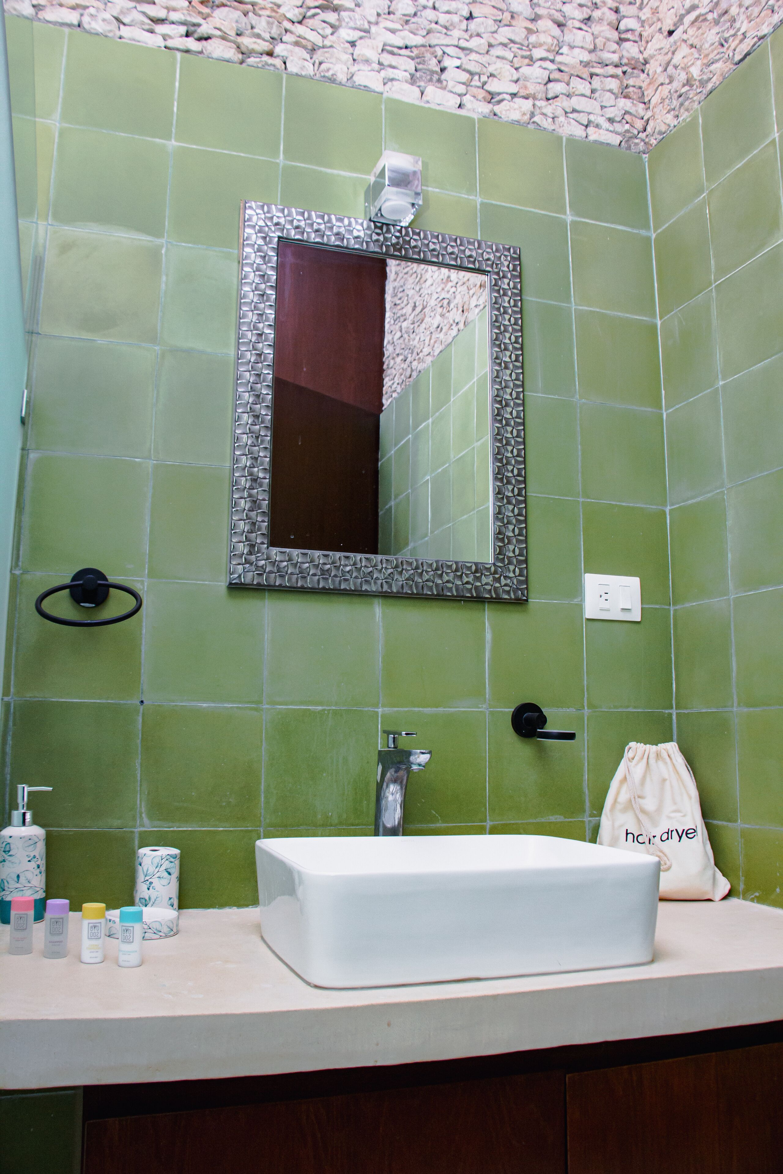 Un baño con azulejos verdes, un lavabo blanco, un espejo con marco decorativo y artículos de aseo en la encimera.