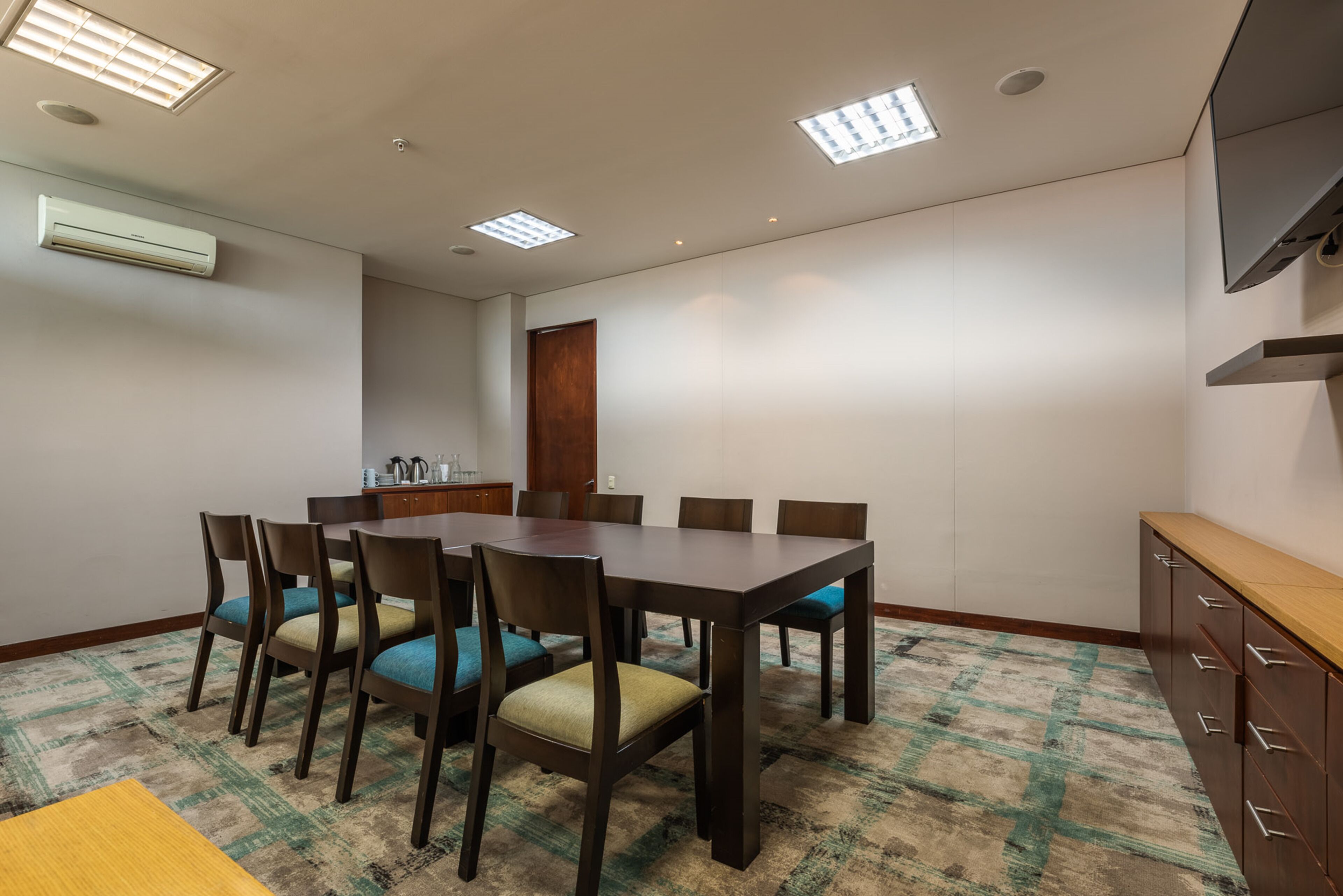 Une salle de conférence avec une table rectangulaire, six chaises, un buffet en bois et une moquette à motifs.