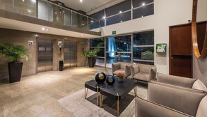 A lobby with a double-height ceiling, a reception desk, two elevators, and a seating area with chairs and a table.