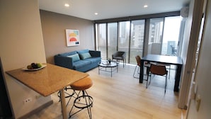 A modern living space with a kitchen area, a dining table with chairs, a sofa, and a large window offering a view of the city.