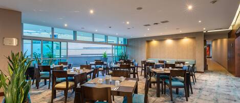 A hotel dining area with multiple tables and chairs, large windows, and indoor plants.