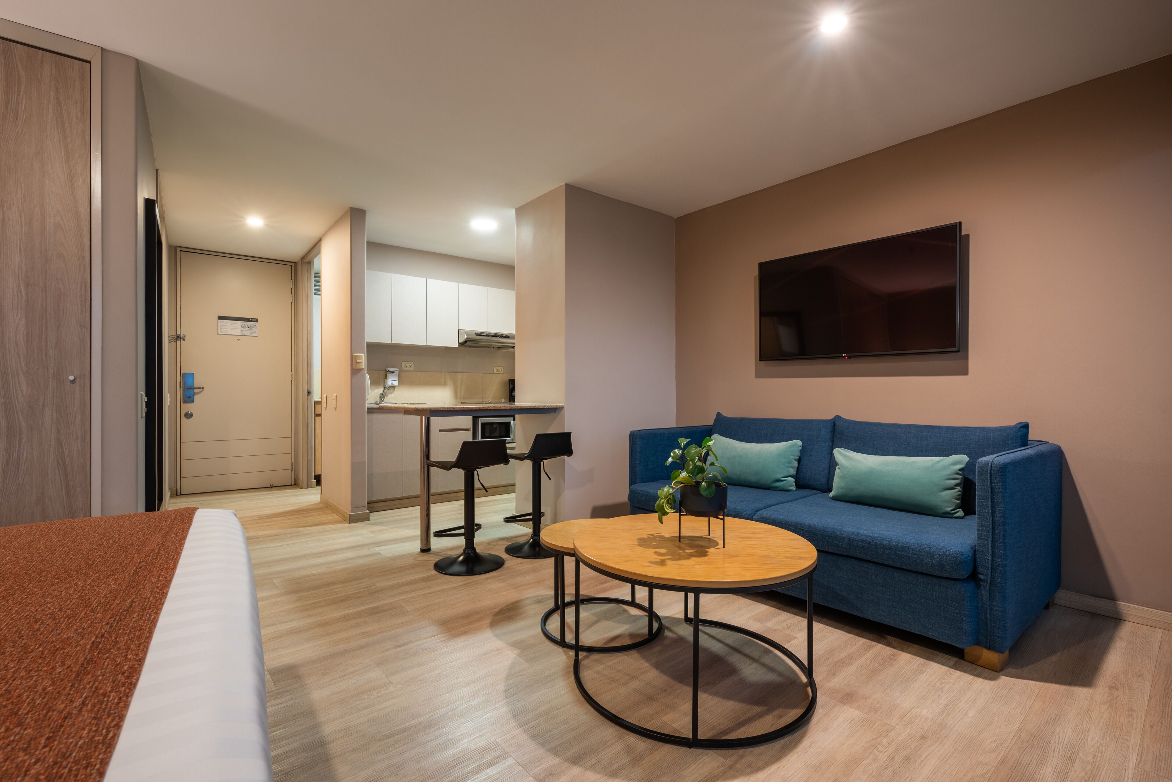 Una habitación de hotel moderna con una cama, una mesa de centro redonda de madera, un sofá azul y una cocina al fondo.