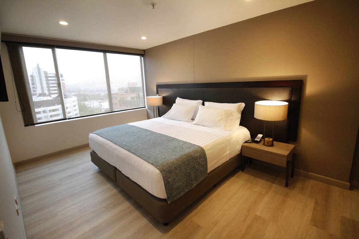 Habitación de hotel moderna con una cama grande, una mesita de noche, una lámpara y vistas a la ciudad a través de amplios ventanales.