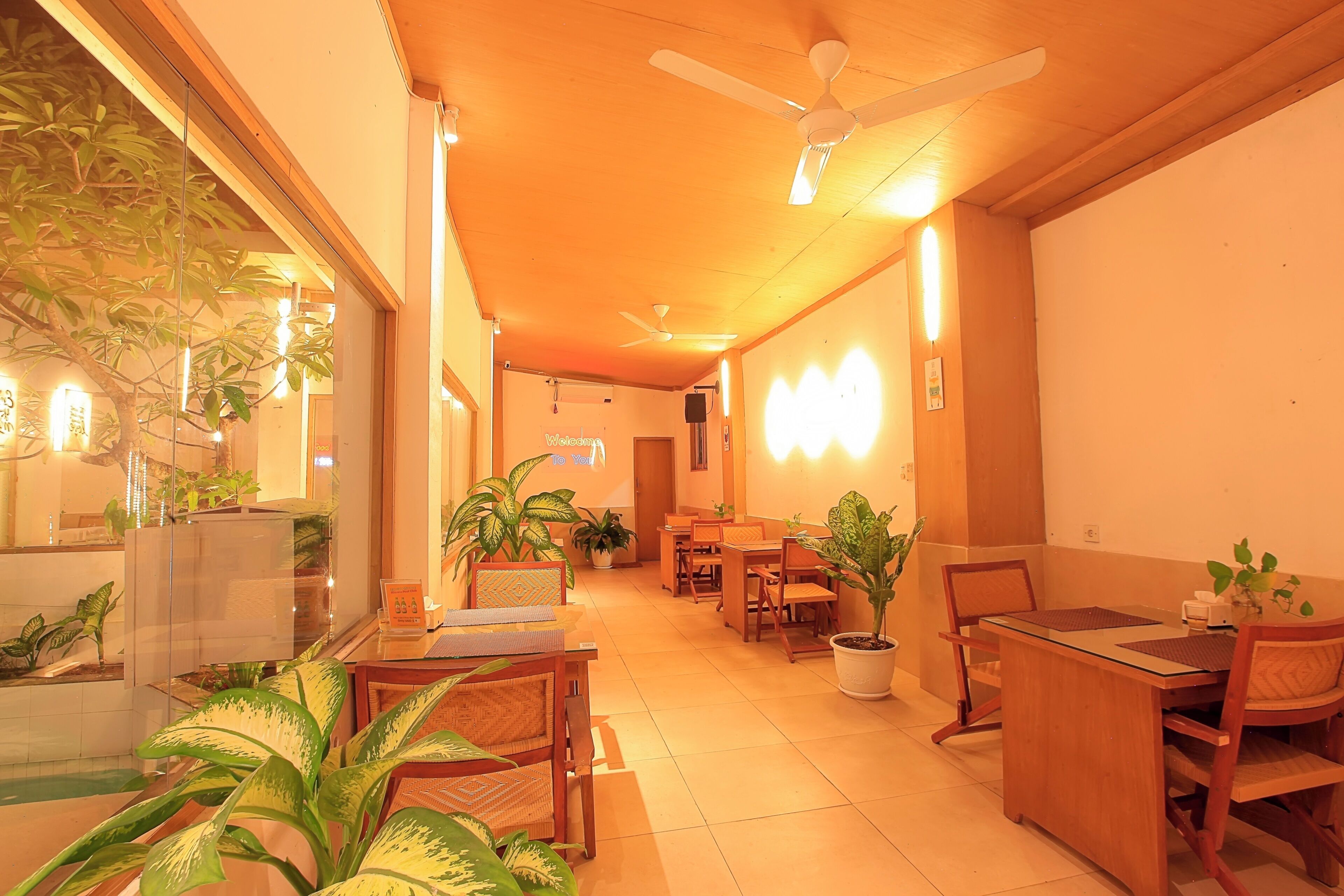 A hotel lobby with a dining area, ceiling fans, potted plants, and a reception desk.