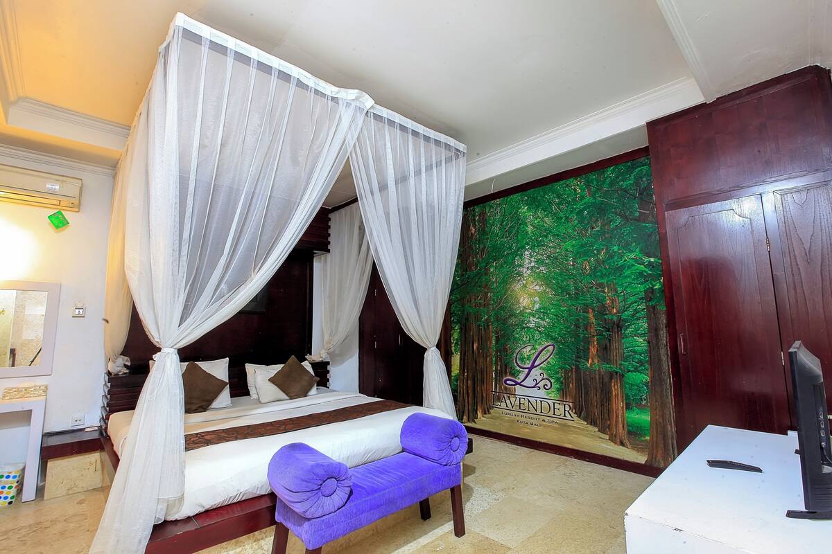 Un lit à baldaquin avec des rideaux blancs, un banc agrémenté d’un coussin violet, un téléviseur posé sur une table blanche et une fresque murale sur le thème de la forêt.