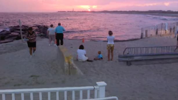 Escena de playa con gente caminando y sentada sobre la arena, un muelle y un atardecer al fondo.