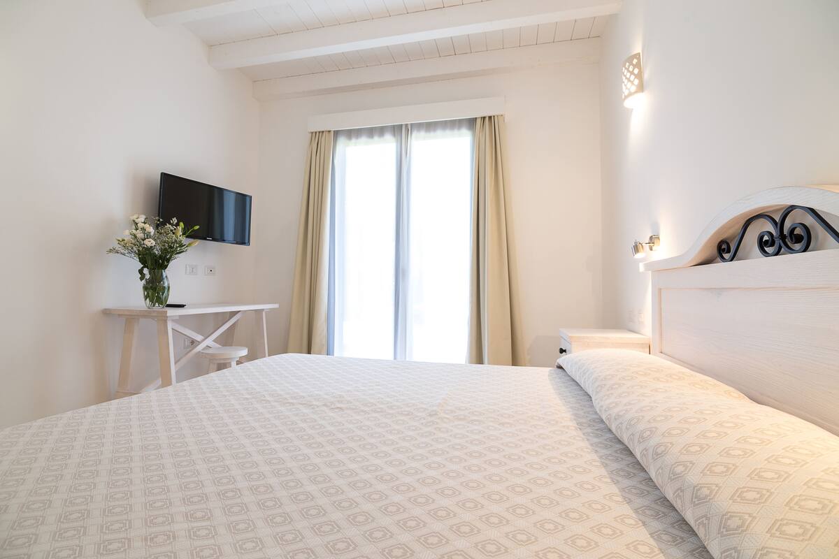 A hotel room with a bed, a TV mounted on the wall, a desk with a vase of flowers, and a window with curtains.