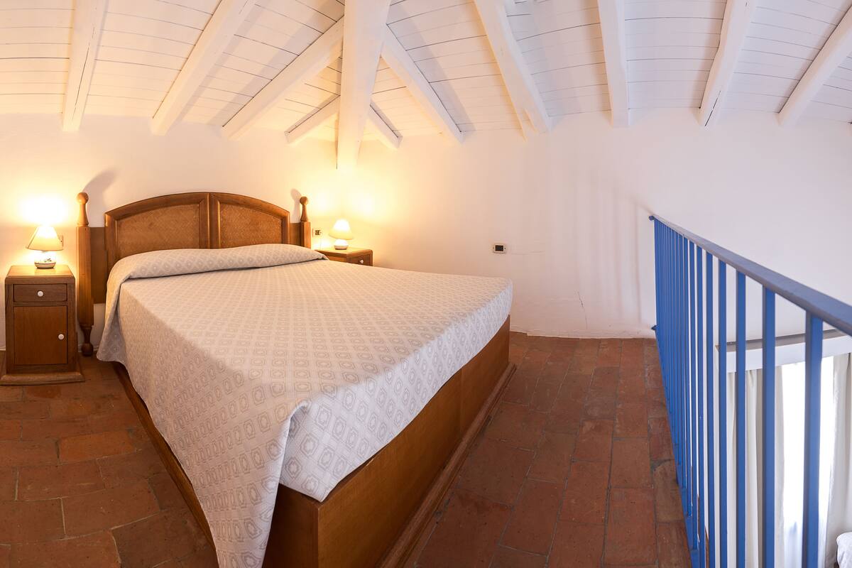 A bedroom with a wooden bed, two bedside lamps, a nightstand, and a view of a blue railing.