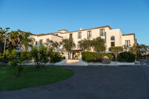 Un hotel de dos pisos con una entrada central, rodeado de jardines bien cuidados y palmeras.