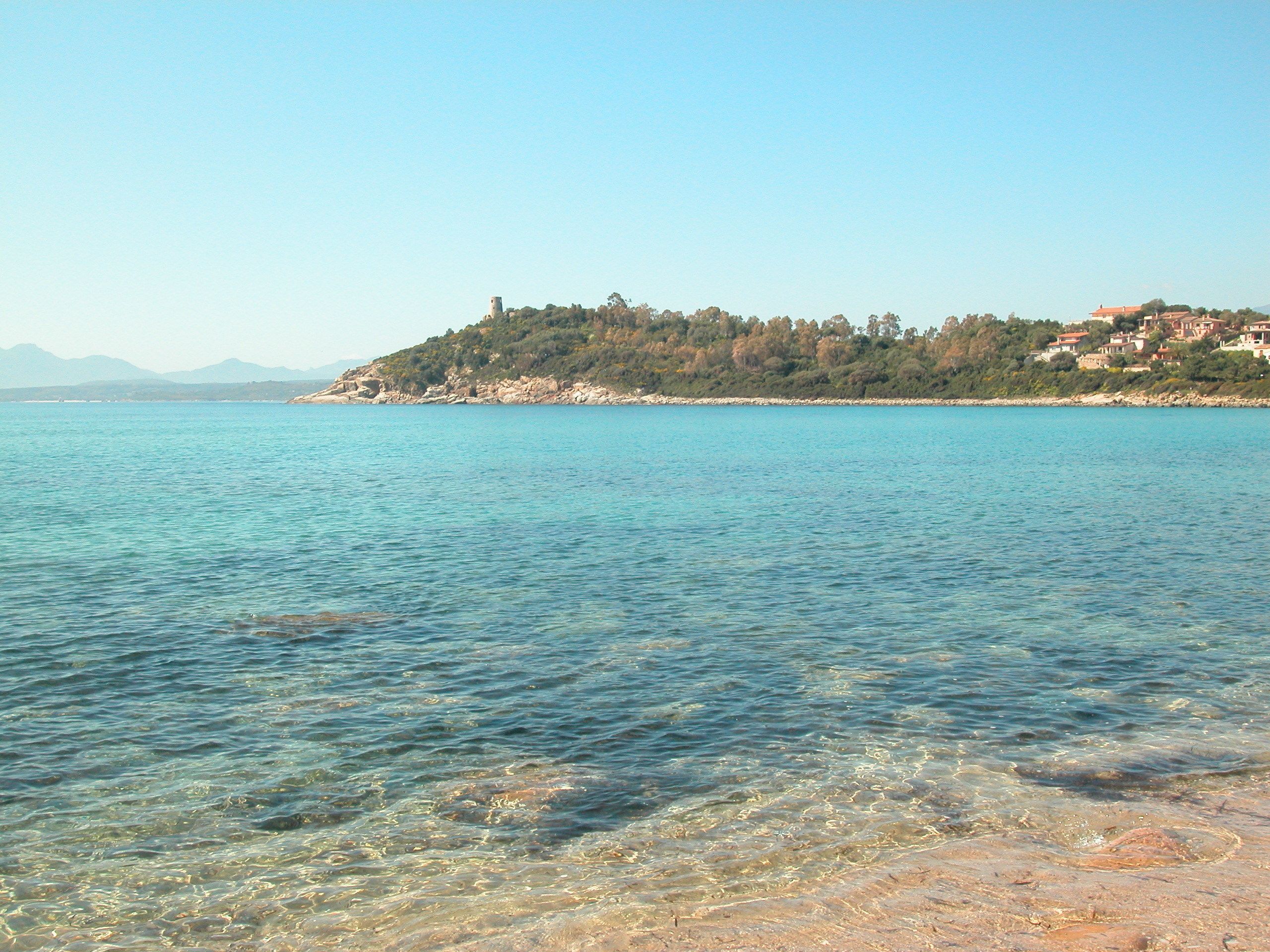 A coastal scene with clear turquoise waters, a rocky shoreline, and a hill with buildings in the background.