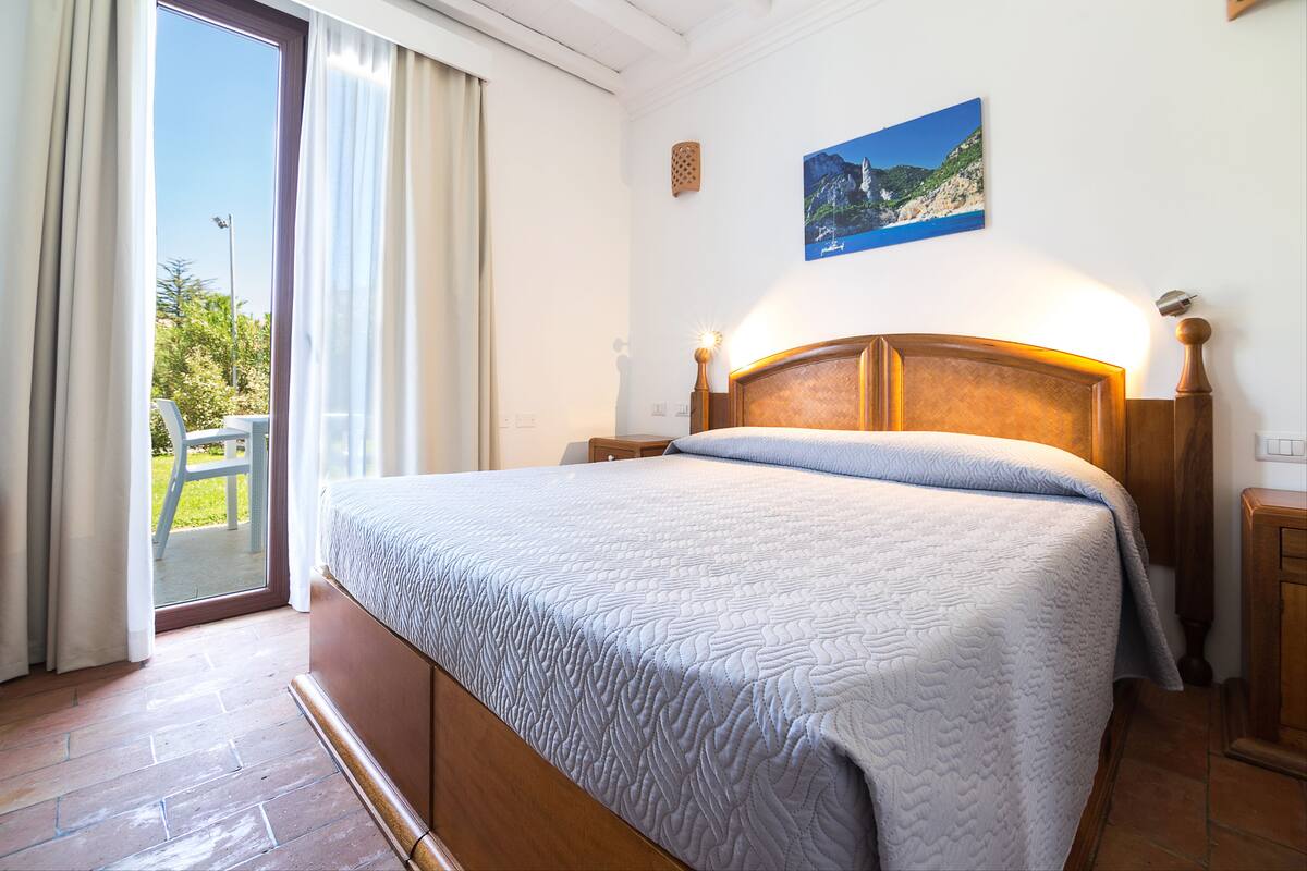 A bedroom with a large bed, a wooden headboard, a nightstand, and a view of the outdoors through a sliding door.