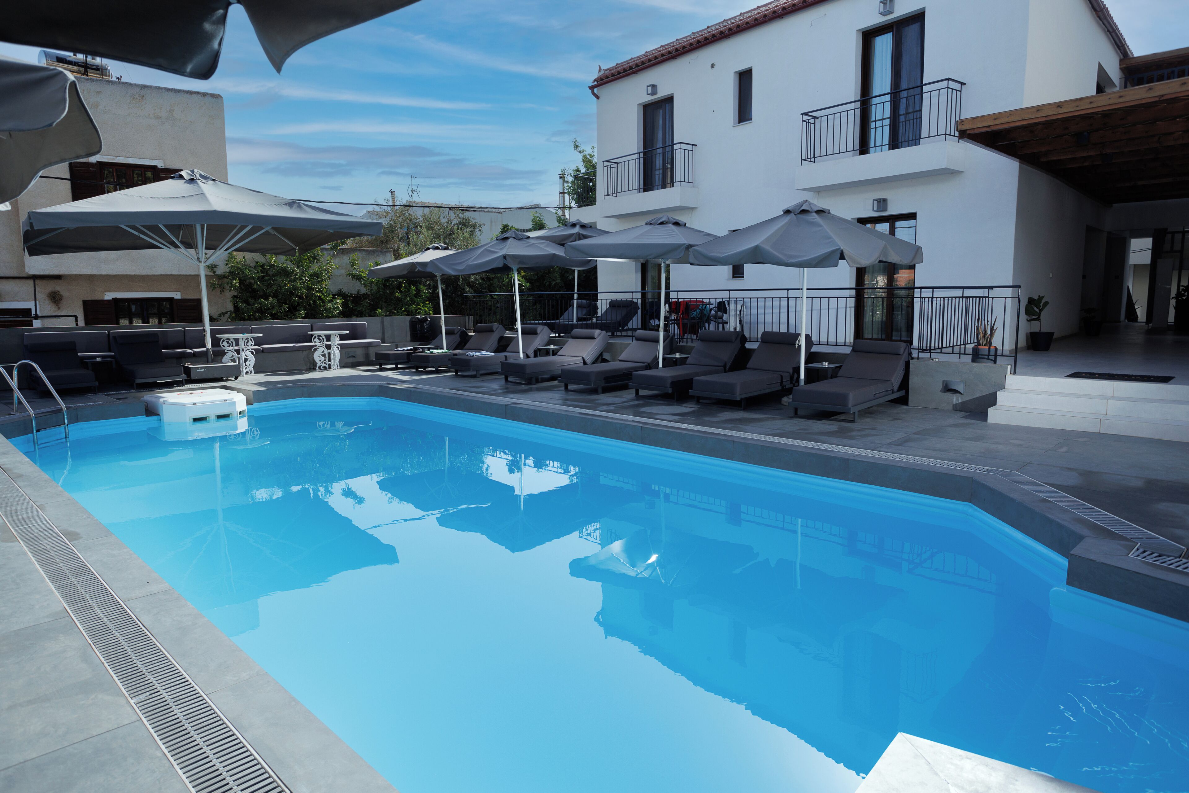 Piscina con sillas de descanso y sombrillas, un edificio blanco con balcones y cielo despejado.