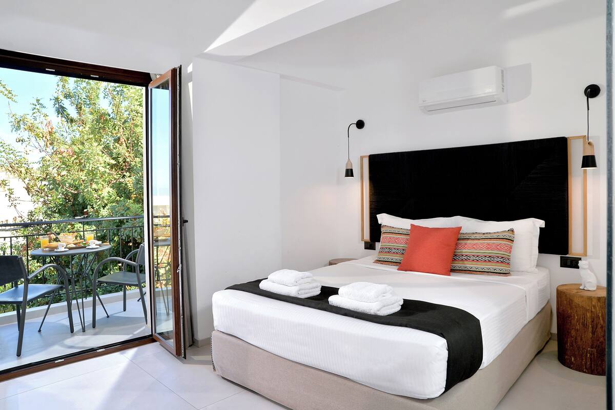 A modern bedroom with a large bed, a balcony with outdoor furniture, and a view of greenery.